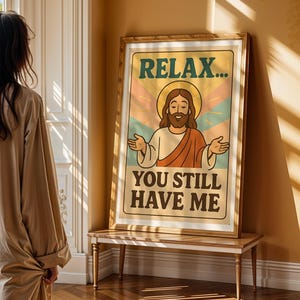 May include: A framed poster with the text "RELAX... YOU STILL HAVE ME" features a cartoon illustration of Jesus with a halo. The poster has a retro color scheme of orange, green, and blue. The poster is displayed on a wooden table.