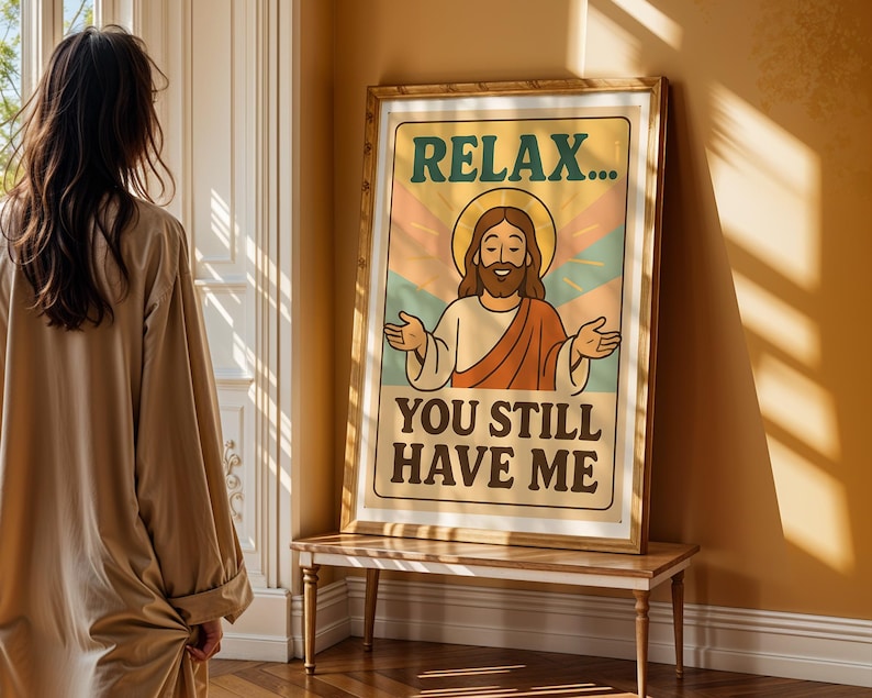 May include: A framed poster with the text "RELAX... YOU STILL HAVE ME" features a cartoon illustration of Jesus with a halo. The poster has a retro color scheme of orange, green, and blue. The poster is displayed on a wooden table.