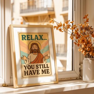 May include: A framed print with the text "RELAX... YOU STILL HAVE ME" featuring an illustration of Jesus with open arms. The artwork has a vintage aesthetic with a beige background and a gold frame. A vase of orange flowers sits nearby.