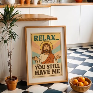 May include: A framed poster with the text "RELAX... YOU STILL HAVE ME" features a cartoon illustration of a figure with a halo. The poster is in a light brown wooden frame. A potted plant and a basket of oranges are also in the image.