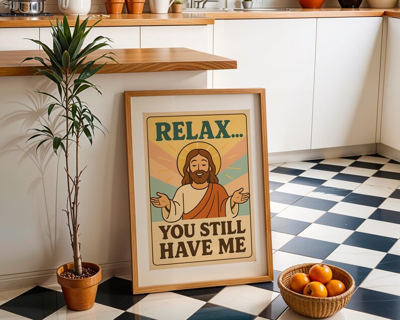 May include: A framed poster with the text "RELAX... YOU STILL HAVE ME" features a cartoon illustration of a figure with a halo. The poster is in a light brown wooden frame. A potted plant and a basket of oranges are also in the image.
