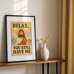 May include: Framed art print featuring a retro-style illustration of Jesus with a halo, arms outstretched. The text reads "RELAX... YOU STILL HAVE ME" in a vintage font. The artwork is in a black frame, displayed on a wooden shelf.