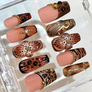 May include: A collection of artificial nails with various designs. Some nails feature a nude base with brown and black animal print patterns, while others have gold embellishments and floral designs. The nails are arranged in a clear plastic container.