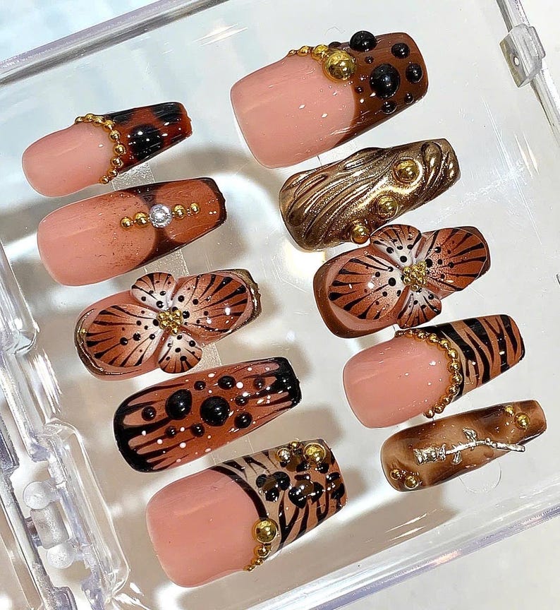 May include: A collection of artificial nails with various designs. Some nails feature a nude base with brown and black animal print patterns, while others have gold embellishments and floral designs. The nails are arranged in a clear plastic container.