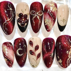 May include: A set of ten almond-shaped press-on nails featuring a deep red magnetic cat-eye finish, cream-colored accents, gold foil flakes, and raised 3D gold embellishments including a bow, a crescent moon, stars, and floral motifs.