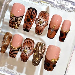 May include: A set of ten artificial nails with various designs. The nails feature a mix of colors, including nude, brown, and gold. Some nails have animal print patterns, floral accents, and decorative embellishments. The nails are arranged in a clear plastic container.