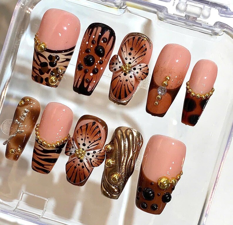 May include: A set of ten artificial nails with various designs. The nails feature a mix of colors, including nude, brown, and gold. Some nails have animal print patterns, floral accents, and decorative embellishments. The nails are arranged in a clear plastic container.