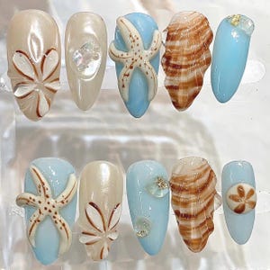 May include: A set of ten artificial nails with a beach theme. The nails feature designs of starfish, seashells, and floral patterns in shades of beige, brown, and blue. Some nails have glitter accents.