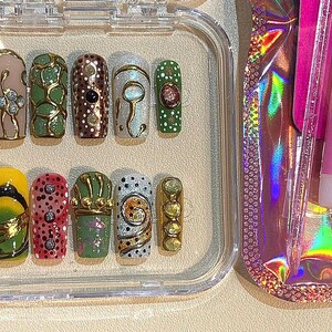 May include: A set of press-on nails in a clear plastic case. The nails feature various designs, including gold accents, dots, and floral patterns. The color palette includes yellow, pink, green, and brown. A pink nail glue and alcohol wipes are visible.