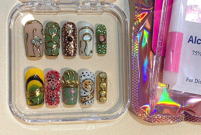 May include: A set of press-on nails in a clear plastic case. The nails feature various designs, including gold accents, dots, and floral patterns. The color palette includes yellow, pink, green, and brown. A pink nail glue and alcohol wipes are visible.