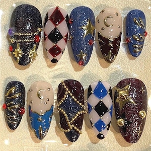 May include: A set of ten artificial nails with various designs. The nails feature glitter, harlequin patterns, stars, moons, and gold embellishments. Colors include navy, red, white, black, and gold. Some nails have a jewel or bead accents.