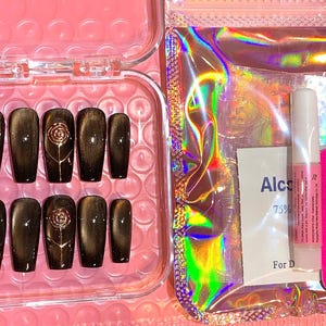 May include: A set of press-on nails in a clear plastic case. The nails are dark brown with a gold rose design. Included are nail glue, a nail file, and an alcohol wipe. The background is a pink surface.