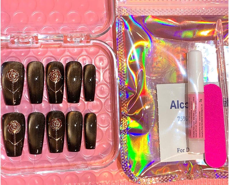 May include: A set of press-on nails in a clear plastic case. The nails are dark brown with a gold rose design. Included are nail glue, a nail file, and an alcohol wipe. The background is a pink surface.