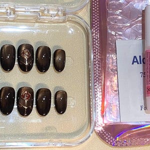 May include: A set of dark brown oval-shaped press-on nails with a rose design. The nails are in a clear plastic case, next to a nail glue tube, a pink nail file, and an alcohol wipe.