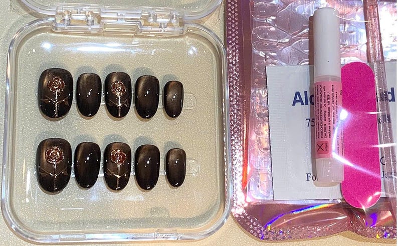 May include: A set of dark brown oval-shaped press-on nails with a rose design. The nails are in a clear plastic case, next to a nail glue tube, a pink nail file, and an alcohol wipe.