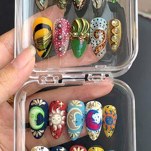 May include: A clear plastic case holds a collection of artificial nails with various designs. The nails feature intricate patterns, including celestial motifs like moons and suns, along with abstract and geometric designs in vibrant colors like green, gold, and blue.