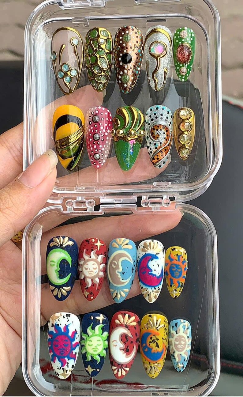 May include: A clear plastic case holds a collection of artificial nails with various designs. The nails feature intricate patterns, including celestial motifs like moons and suns, along with abstract and geometric designs in vibrant colors like green, gold, and blue.