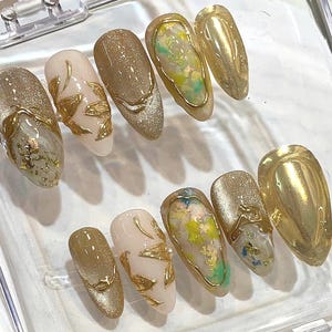 May include: A set of ten artificial nails in various designs. Some nails are gold glitter, others are a light beige with gold leaf accents. Several nails feature a gold border with colorful accents.