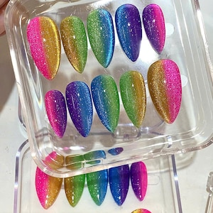 May include: A set of colorful, glittery false nails in a clear plastic case. The nails are in a variety of colors, including pink, gold, green, blue, and purple, with a gradient effect. The nails are almond-shaped.
