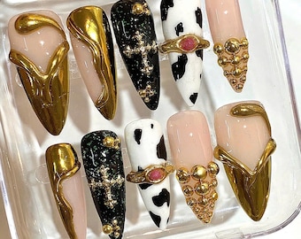 May include: A set of ten artificial nails in various designs. Some nails are nude with gold accents, others are black with gold crosses, and some have a cow print design. The nails are in a clear plastic case.