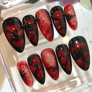 May include: A set of ten artificial nails with a glossy finish. Each nail features a blend of black and red hues, creating a marbled effect. The nails are oval-shaped and arranged in a clear plastic container.