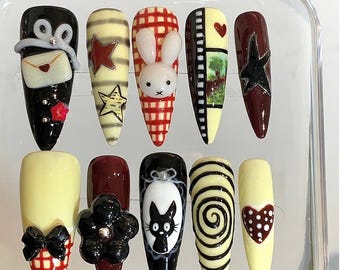May include: A collection of ten artificial nails with diverse designs. The nails showcase colours like black, red, yellow, and white, with decorative elements such as stars, bows, a bunny, and a cat. The nails are presented in a clear plastic case.