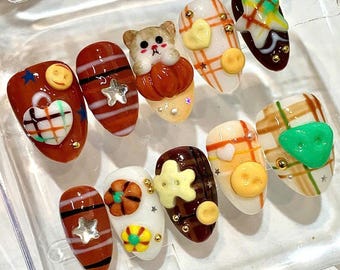 May include: A set of ten press-on nails featuring various designs including plaid patterns, 3D bear and pumpkin figures, smiley faces, stars, hearts, and flowers in a color palette of brown, white, orange, yellow, and green with gold accents.