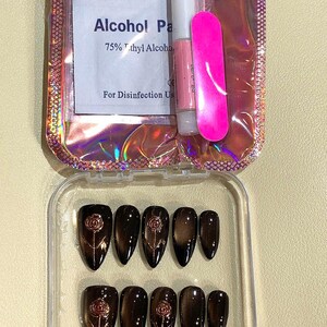 May include: A set of press-on nails in a clear plastic case. The nails are dark brown with a rose design. Included are nail glue, a pink nail file, and an alcohol wipe. The packaging is iridescent.