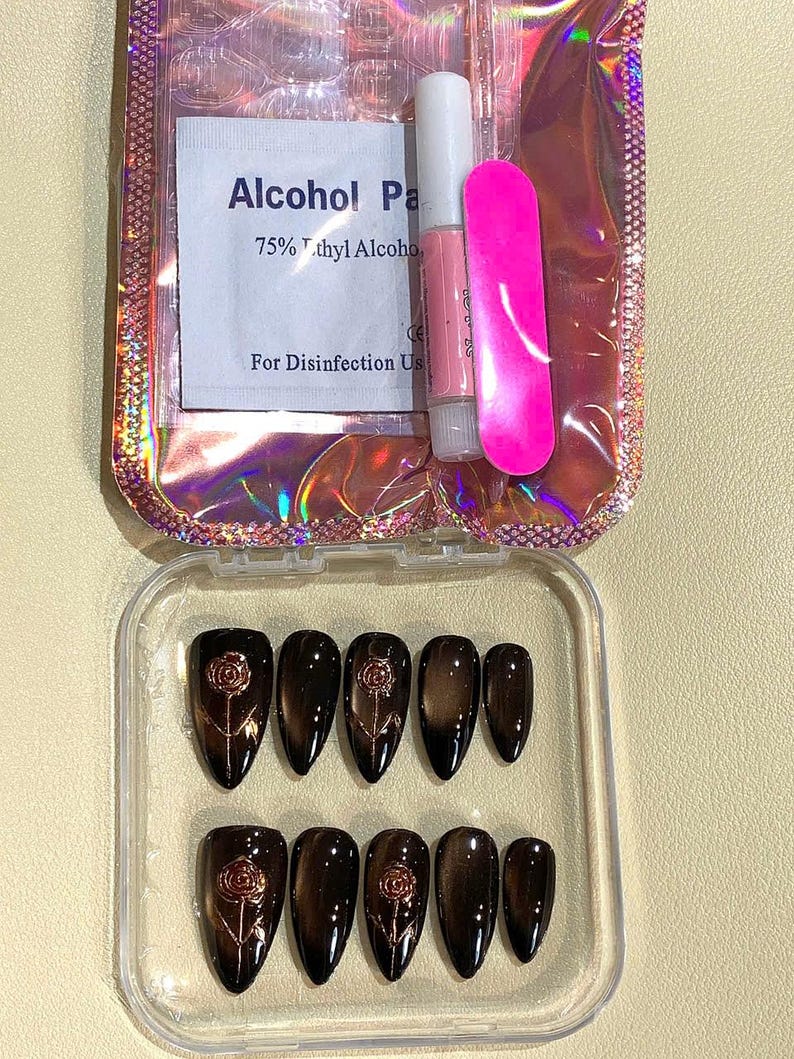 May include: A set of press-on nails in a clear plastic case. The nails are dark brown with a rose design. Included are nail glue, a pink nail file, and an alcohol wipe. The packaging is iridescent.