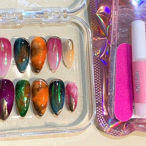 Puede incluir: Un juego de u&ntilde;as postizas ovaladas en una caja de pl&aacute;stico transparente. Las u&ntilde;as presentan una variedad de colores, incluyendo rosa, verde, naranja y dorado. Tambi&eacute;n se incluyen una lima de u&ntilde;as rosa, pegamento para u&ntilde;as y una almohadilla con alcohol.