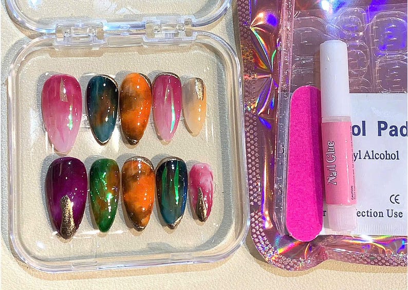 Puede incluir: Un juego de u&ntilde;as postizas ovaladas en una caja de pl&aacute;stico transparente. Las u&ntilde;as presentan una variedad de colores, incluyendo rosa, verde, naranja y dorado. Tambi&eacute;n se incluyen una lima de u&ntilde;as rosa, pegamento para u&ntilde;as y una almohadilla con alcohol.