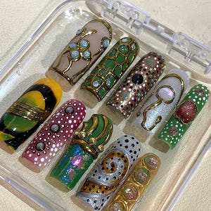 May include: A set of ten artificial nails in a clear plastic case. The nails feature various designs, including floral patterns, geometric shapes, and abstract art. Colors include gold, green, pink, and white. Each nail is unique, showcasing intricate details and embellishments.