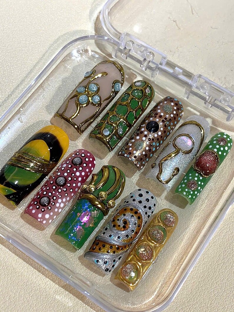 May include: A set of ten artificial nails in a clear plastic case. The nails feature various designs, including floral patterns, geometric shapes, and abstract art. Colors include gold, green, pink, and white. Each nail is unique, showcasing intricate details and embellishments.