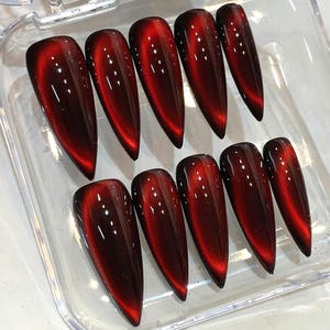 May include: A collection of ten long, pointed false nails in a deep red hue with a cat-eye effect. The nails are arranged in two rows of five, with a glossy finish and a subtle shimmer.