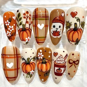 Op de afbeelding: Een set van tien amandelvormige kunstnagels met herfstthema, waaronder oranje pompoenen, witte bloemen, bruine ruitpatronen, een klein spookje met een muts, een hartje, een koffiemok met slagroom en een bruine strik.