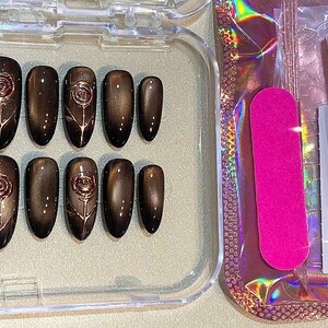 May include: A set of press-on nails in a clear plastic case. The nails are a dark brown color with a rose design. A pink nail file and nail glue are also included.