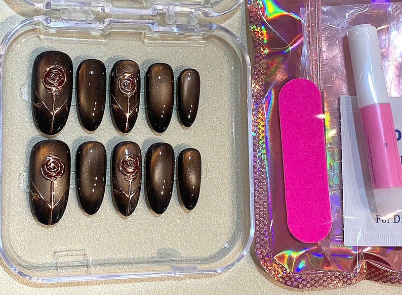 May include: A set of press-on nails in a clear plastic case. The nails are a dark brown color with a rose design. A pink nail file and nail glue are also included.