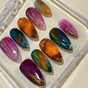 Puede incluir: Un juego de diez u&ntilde;as postizas en forma de almendra en una caja de pl&aacute;stico transparente. Las u&ntilde;as presentan una variedad de colores, incluyendo rosa, naranja, azul, verde y morado, con detalles met&aacute;licos dorados. Las u&ntilde;as tienen un acabado brillante.