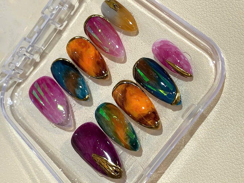 Puede incluir: Un juego de diez u&ntilde;as postizas en forma de almendra en una caja de pl&aacute;stico transparente. Las u&ntilde;as presentan una variedad de colores, incluyendo rosa, naranja, azul, verde y morado, con detalles met&aacute;licos dorados. Las u&ntilde;as tienen un acabado brillante.