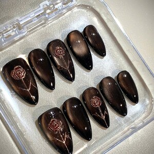 May include: Set of ten dark brown, almond-shaped false nails with a glossy finish. Each nail features a rose design in a lighter brown color. The nails are arranged in a clear plastic container.
