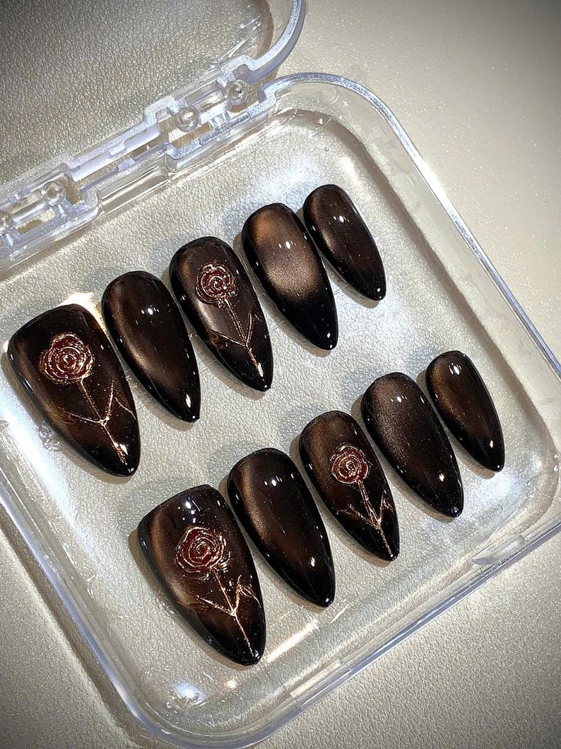 May include: Set of ten dark brown, almond-shaped false nails with a glossy finish. Each nail features a rose design in a lighter brown color. The nails are arranged in a clear plastic container.