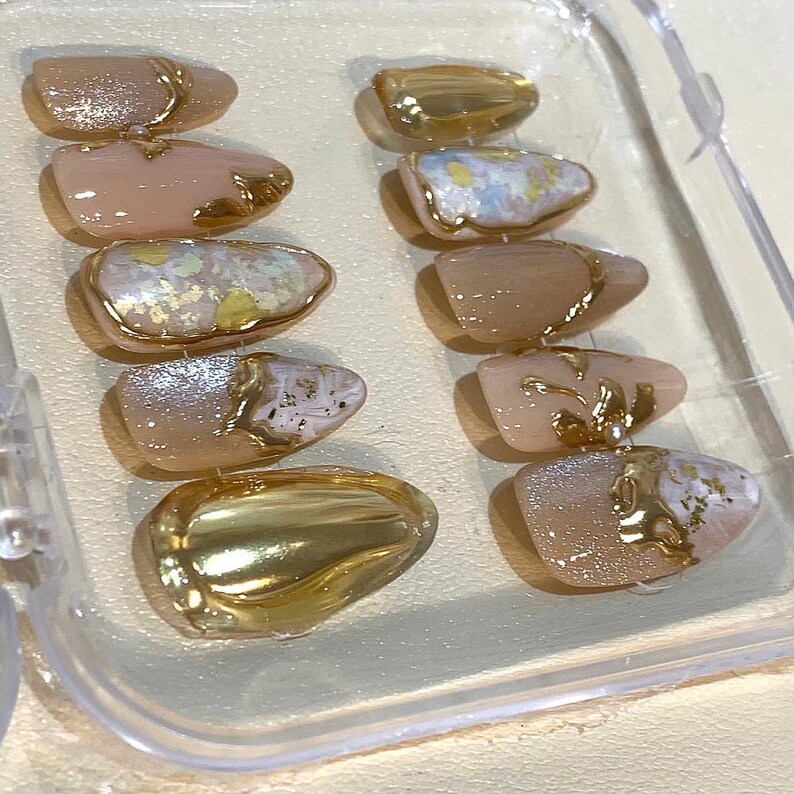 May include: A set of ten artificial nails in a clear plastic container. The nails feature a variety of designs, including gold metallic accents, glitter, and iridescent finishes. The base colors are primarily nude and pale pink.