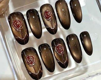 May include: Set of ten oval-shaped, dark brown press-on nails. Each nail has a glossy finish and features a red rose design with gold accents. The nails are arranged in a clear plastic container, showcasing a manicure set.