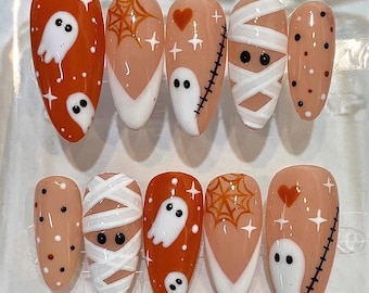 May include: A set of ten almond-shaped press-on nails featuring Halloween designs. The nails include orange backgrounds with white ghosts, nude bases with orange spider webs, mummy bandages, black stitches, red hearts, and black and white polka dots.