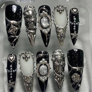 Puede incluir: Un juego de diez uñas postizas en forma de estilete con una paleta de colores en blanco y negro. Los diseños incluyen emblemas de cruces de metal plateado, marcos ovalados adornados con centros brillantes, corazones, estrellas y perlas.