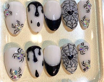 May include: A set of Halloween-themed false nails. The nails feature designs including spiderwebs, spiders, and dripping black paint. Some nails have a black and silver French tip. Embellishments include iridescent rhinestones.