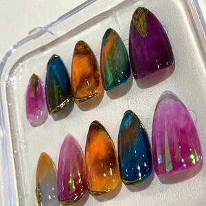 Puede incluir: Un juego de diez u&ntilde;as artificiales en una caja de pl&aacute;stico transparente. Las u&ntilde;as presentan una variedad de colores, incluyendo morado, naranja, azul y rosa, con detalles dorados. Cada u&ntilde;a tiene un acabado brillante.