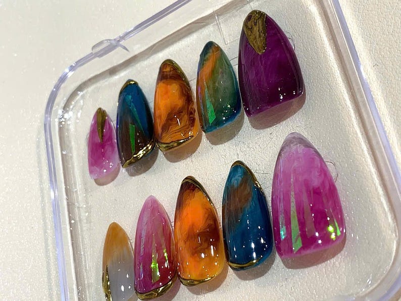 Puede incluir: Un juego de diez u&ntilde;as artificiales en una caja de pl&aacute;stico transparente. Las u&ntilde;as presentan una variedad de colores, incluyendo morado, naranja, azul y rosa, con detalles dorados. Cada u&ntilde;a tiene un acabado brillante.