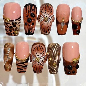 May include: A set of ten artificial nails in various designs. The nails feature a combination of nude, brown, and gold colors. Some nails have animal print patterns, floral designs, and gold embellishments. The nails are arranged in a clear plastic container.