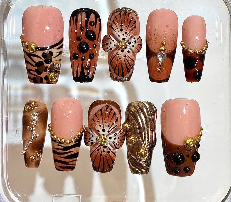May include: A set of ten artificial nails in various designs. The nails feature a combination of nude, brown, and gold colors. Some nails have animal print patterns, floral designs, and gold embellishments. The nails are arranged in a clear plastic container.